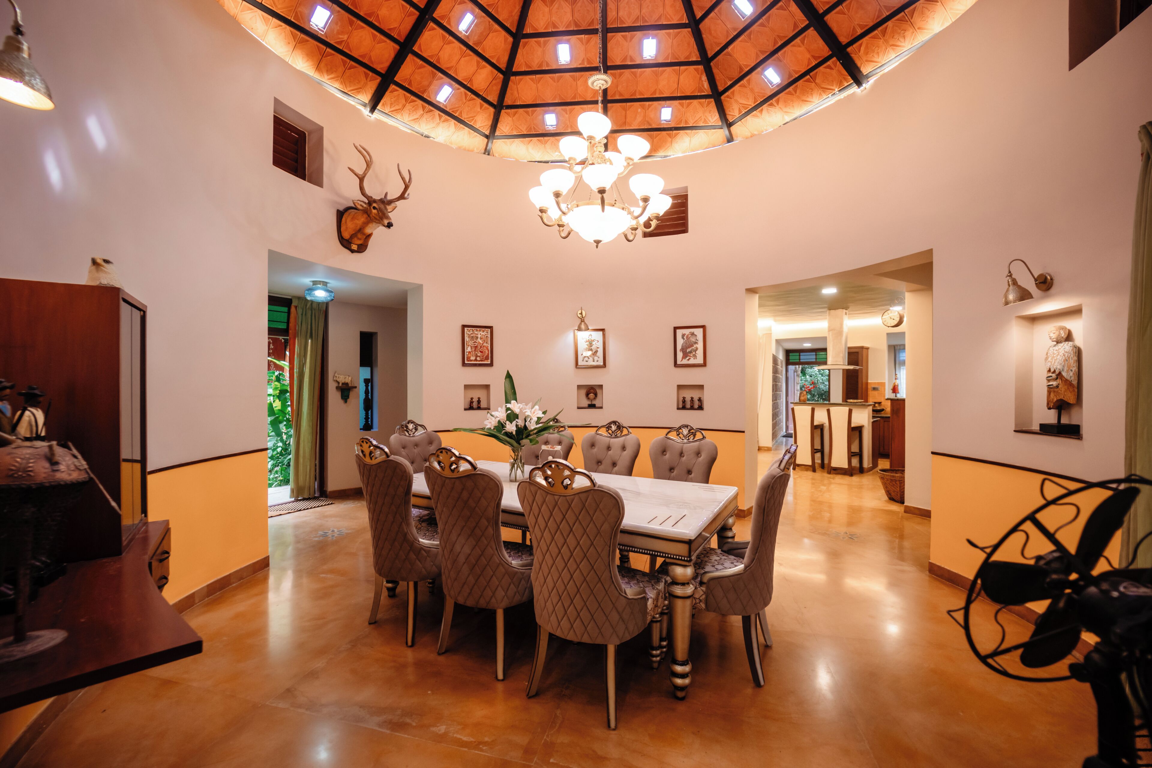 A dining area with a round table, multiple chairs, a chandelier, and a ceiling with a geometric pattern.
