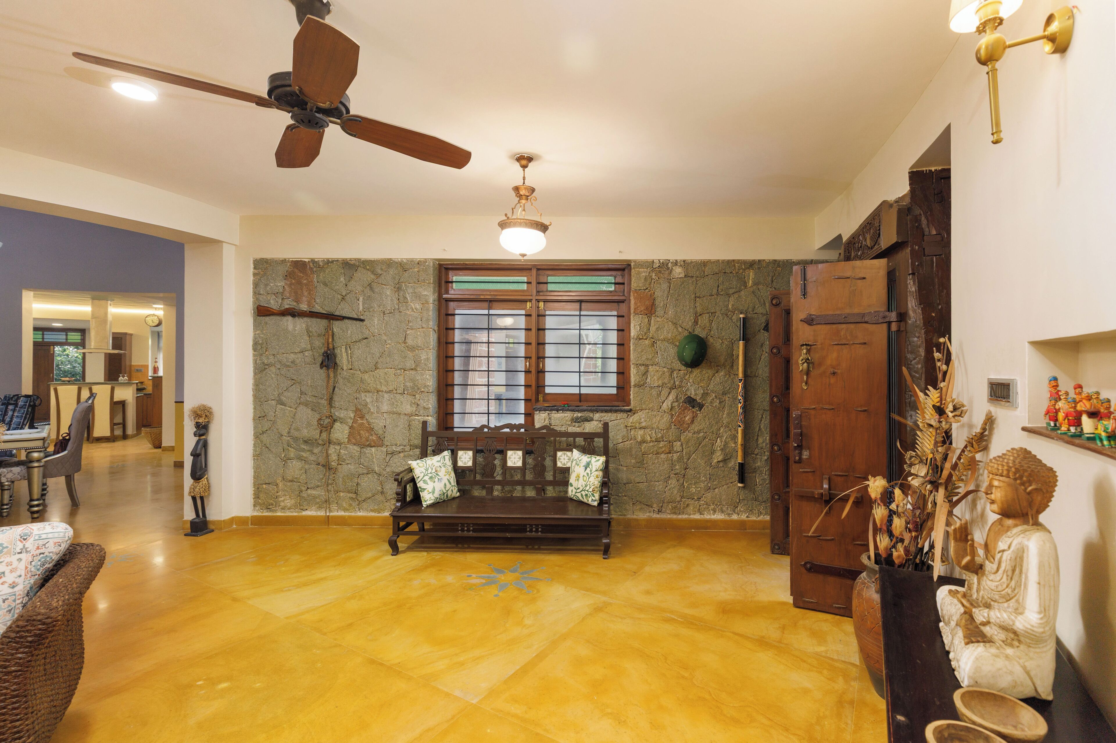 Un salón amplio con un banco de madera, un ventilador de techo, una pared de piedra y un ventanal grande.