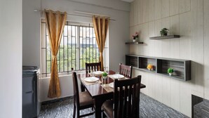 Un coin repas avec une table dressée pour deux personnes, des chaises, une fenêtre avec des rideaux et des étagères intégrées ornées de plantes.