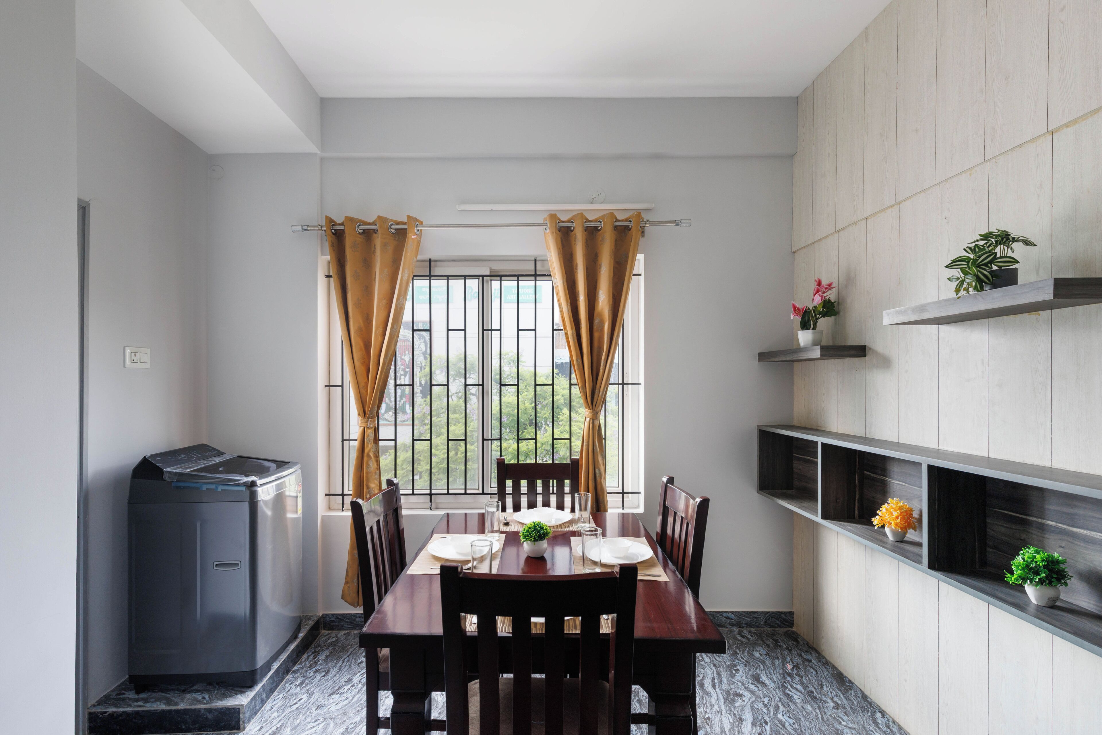 A dining area with a table set for two, a washing machine, and a view through a window with curtains.