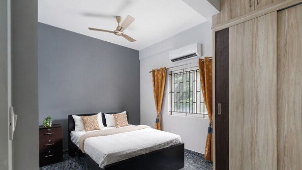 Une chambre à coucher comprenant un lit, une table de chevet, une armoire, un ventilateur de plafond et une fenêtre avec des rideaux.