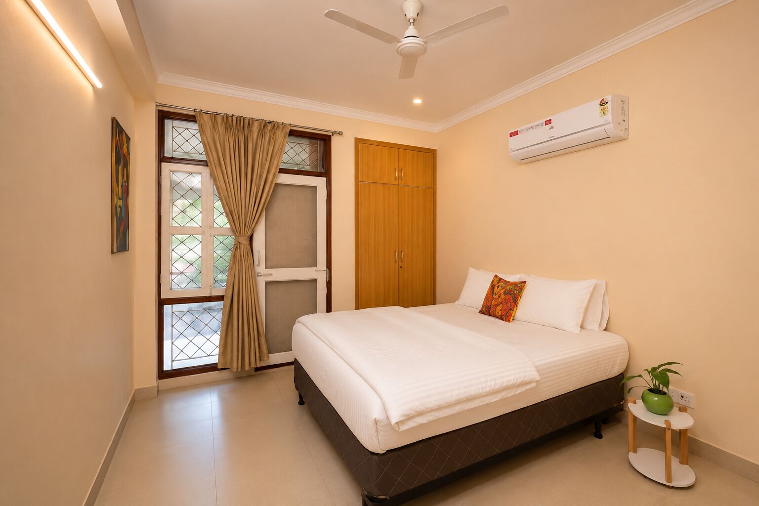 A bedroom with a bed, a ceiling fan, an air conditioning unit, a window with curtains, and a small plant on a side table.