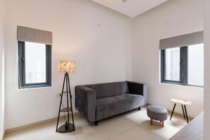 A modern living room with a grey sofa, a floor lamp, a small round ottoman, and two windows with blinds. - Orionrosewood Villa by Redolive3bhkacnr Airport (Bengaluru)