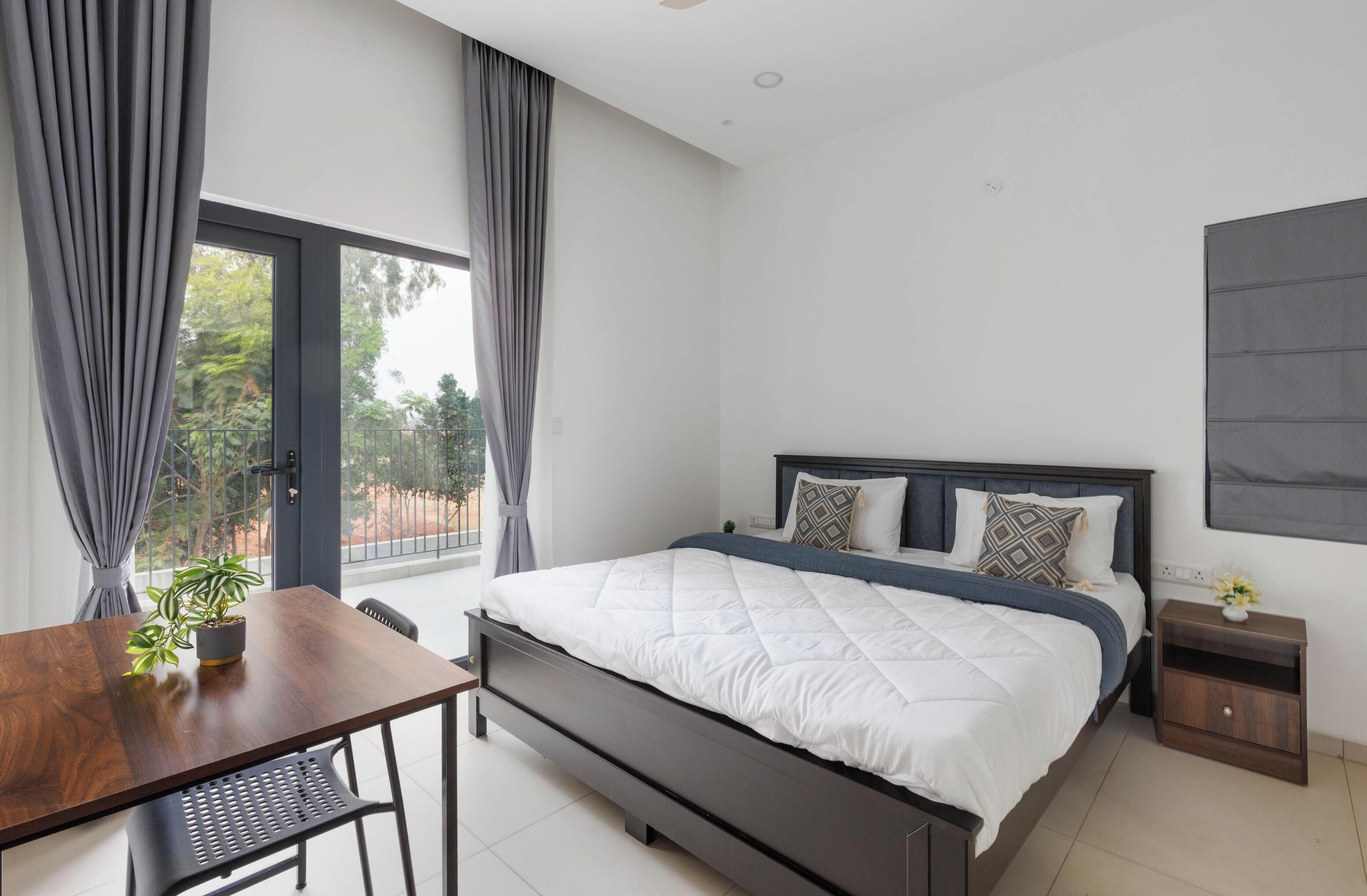 A modern bedroom with a large bed, a wooden desk, and a view of the outdoors through a sliding door.