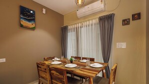 A dining area with a wooden table set for four, chairs, a painting of a beach scene, an air conditioner, and a window with curtains. - Orionrosewood Villa by Redolive3bhkacnr Airport (Bengaluru)