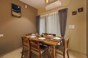 A dining area with a wooden table set for four, chairs, a painting of a beach scene, an air conditioner, and a window with curtains. - Orionrosewood Villa by Redolive3bhkacnr Airport (Bengaluru)
