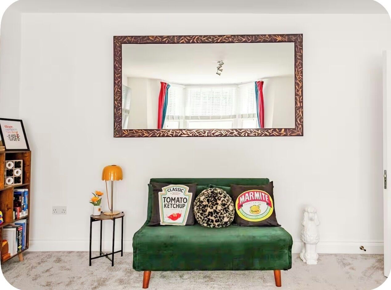 Une pièce avec un canapé en velours vert, une petite table d’appoint avec une lampe, une bibliothèque et un grand miroir doté d’un cadre à motifs.