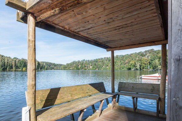 Une jetée en bois avec un banc, d’où l’on peut admirer un lac paisible, avec une colline boisée en arrière-plan.