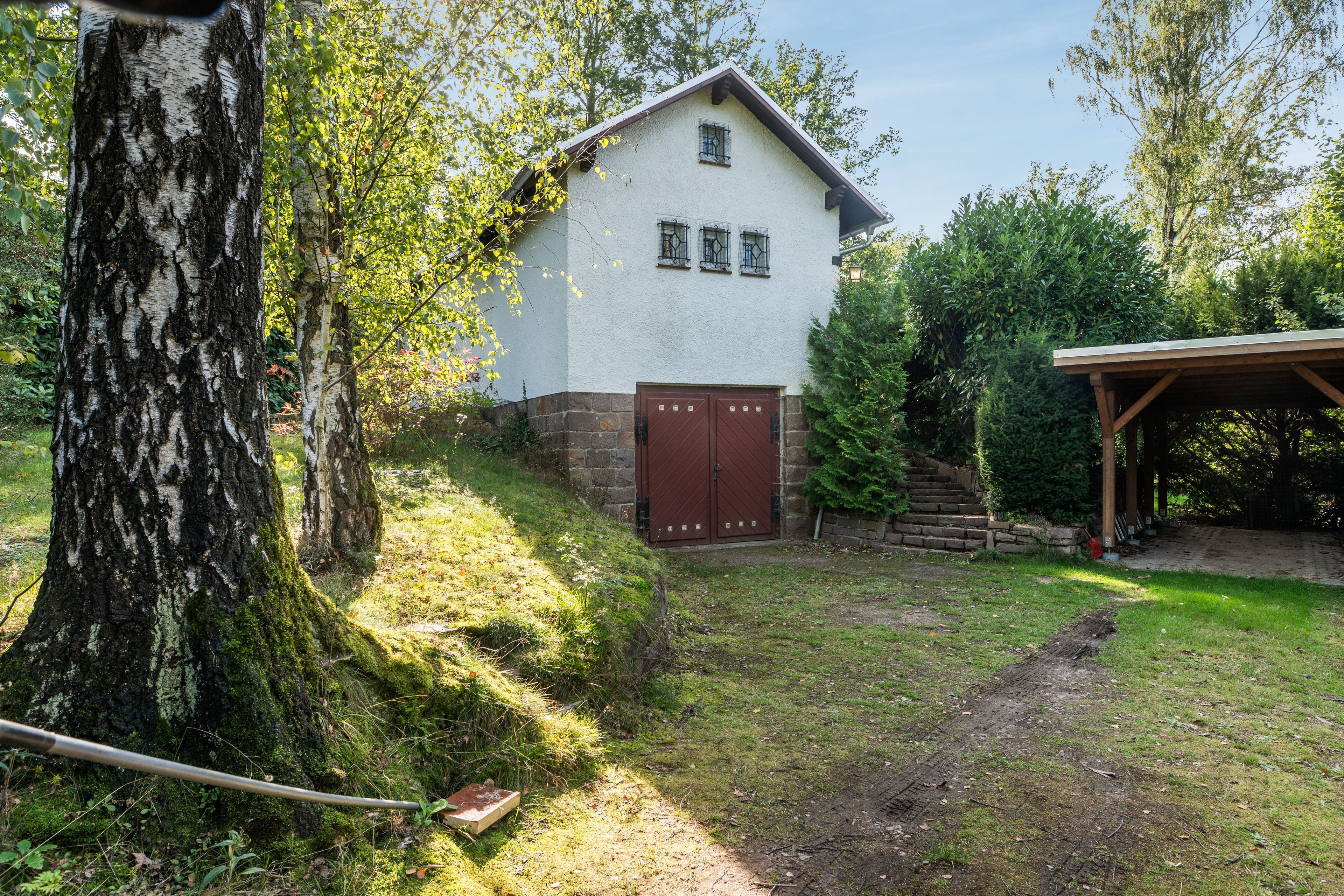Ein Gebäude mit weißer Fassade und brauner Tür, umgeben von Grünpflanzen und einem überdachten Außenbereich.