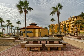 A courtyard with a central table and benches, surrounded by palm trees and a building with a yellow facade. - Princesa Resort A-506 Family Tides Refuge Pleasant Ocean Front Condo (Puerto Peñasco)