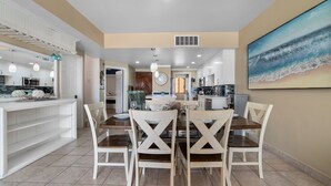 Una cucina moderna con mobili bianchi, un'area pranzo con tavolo di legno e sedie bianche, e un quadro a tema spiaggia al muro.