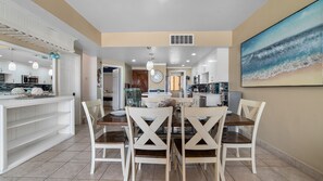 A modern kitchen with white cabinets, a dining area with a wooden table and white chairs, and a beach-themed painting on the wall. - Princesa Resort A-506 Family Tides Refuge Pleasant Ocean Front Condo (Puerto Peñasco)
