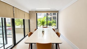 A conference room with a long table and chairs, large windows with blinds, and a view of greenery outside.