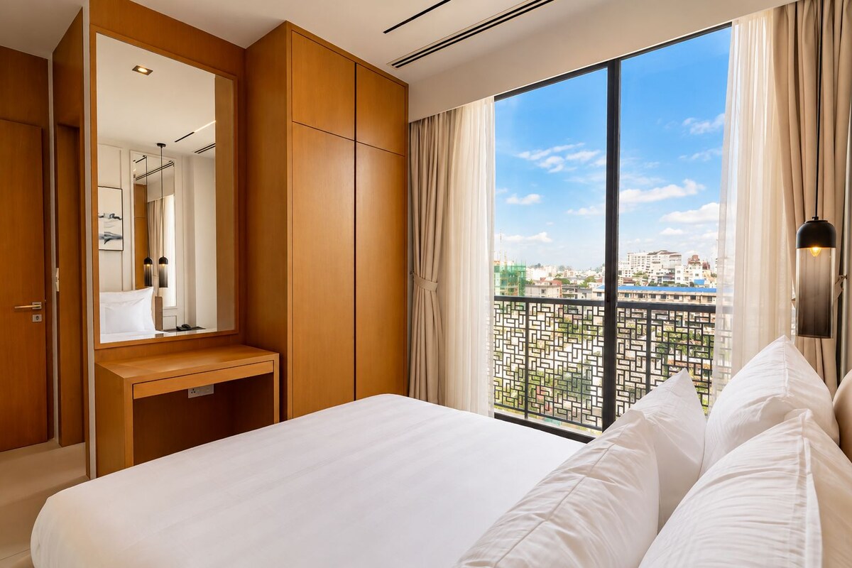 Habitación de hotel con una cama grande, un armario de madera, una mesita de noche, un espejo y vistas a la ciudad desde un balcón.