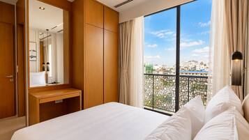 A hotel room with a large bed, a wooden wardrobe, a bedside table, a mirror, and a view of the city through a balcony.