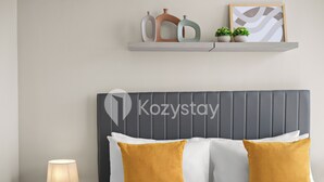A hotel room with a grey upholstered headboard, a bed with white and mustard-colored pillows, a bedside table with a lamp, and a shelf with decorative items and plants. - Corvia by Kozystay - Kemayoran (Jakarta Pusat)