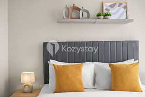 A hotel room with a grey upholstered headboard, a bed with white and mustard-colored pillows, a bedside table with a lamp, and a shelf with decorative items and plants. - Corvia by Kozystay - Kemayoran (Jakarta Pusat)