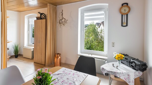 Een kamer met een houten plafond, een raam met uitzicht op groen, een wandklok en een tafel met een bloemrijk tafelkleed.