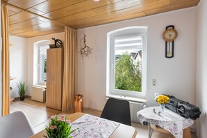 A room with a wooden ceiling, a window with a view of greenery, a wall clock, and a table with a floral tablecloth.