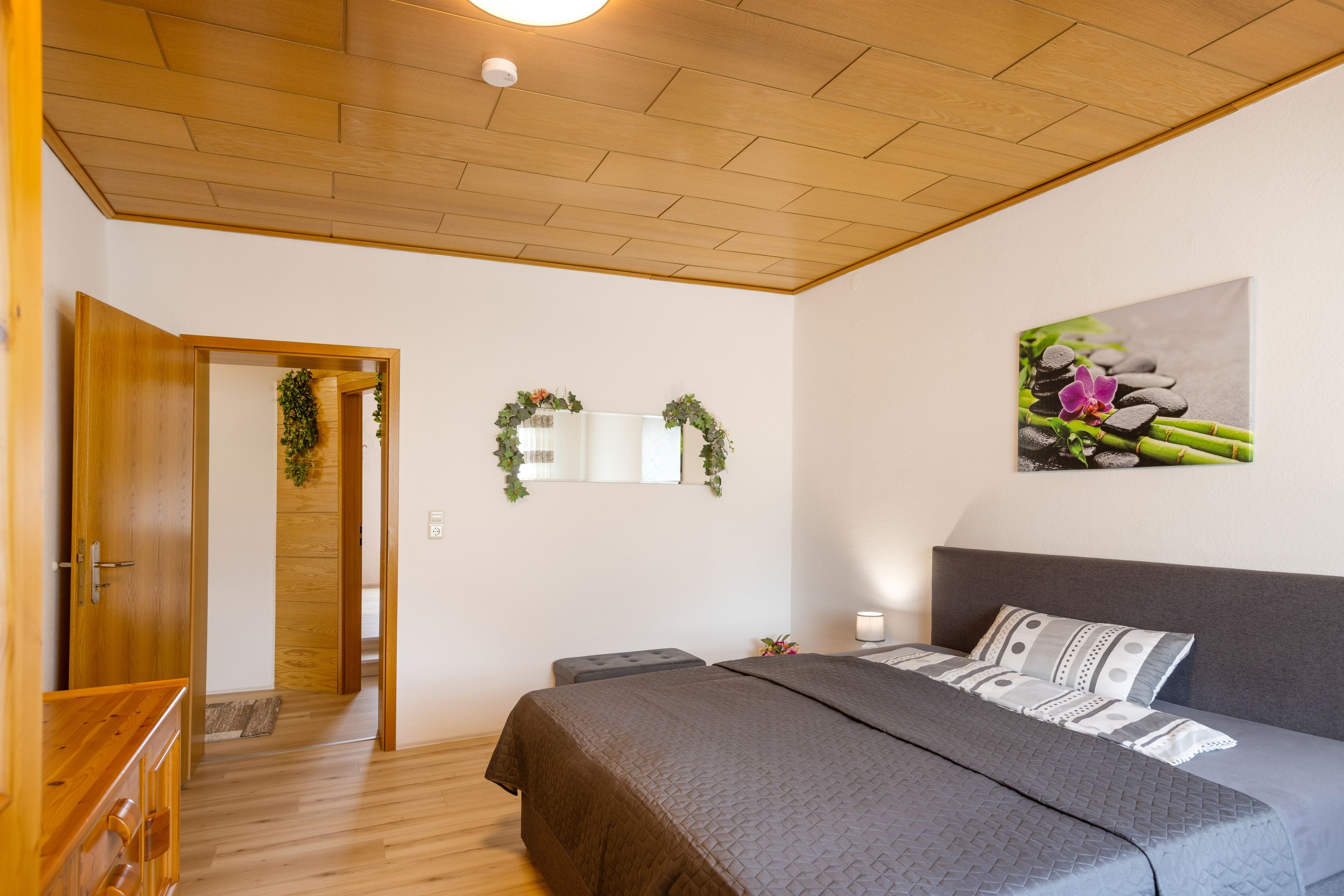 Un dormitorio con techo de madera, una cama con cabecera gris, una mesita de noche y un cuadro colgado en la pared.