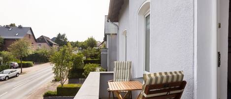 Ein Balkon mit Tisch und zwei Stühlen, der auf eine Straße mit parkenden Autos und Häusern blickt.