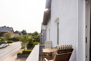 A balcony with a table and two chairs, overlooking a street with parked cars and houses.