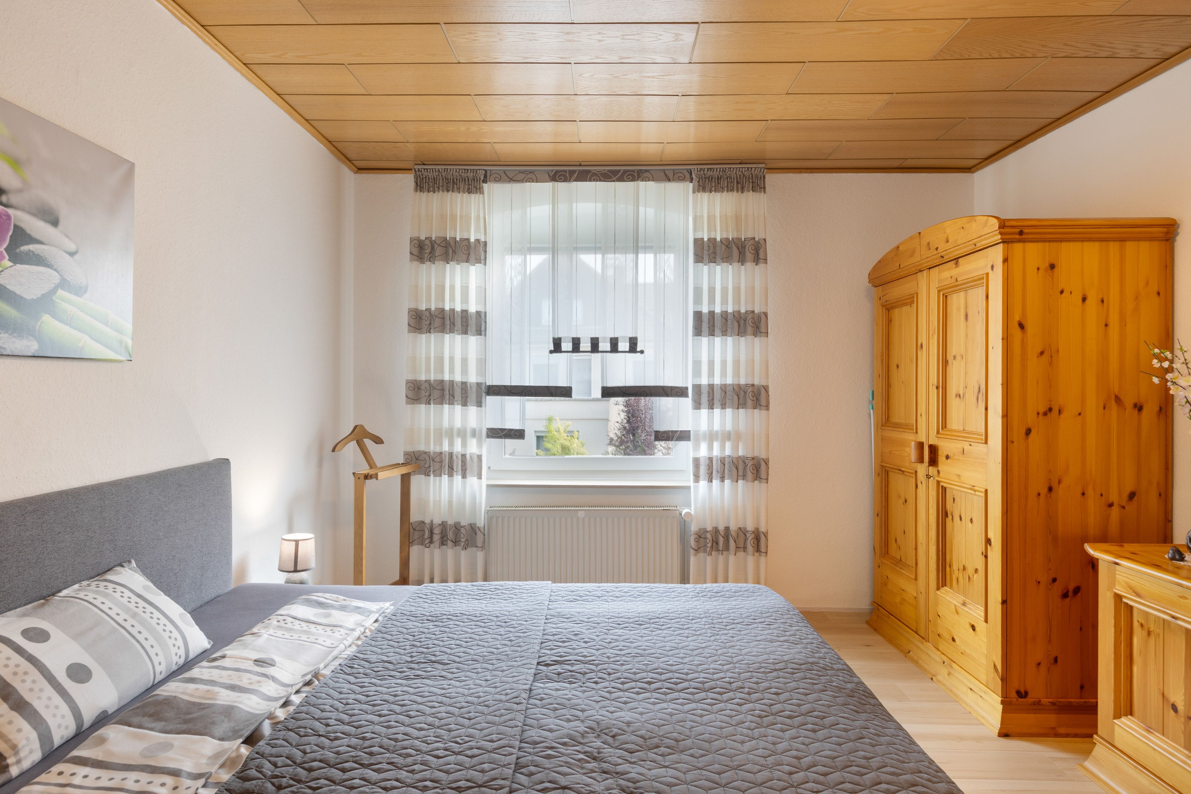 Quarto com cama, guarda-roupa de madeira, radiador e janela com cortinas.