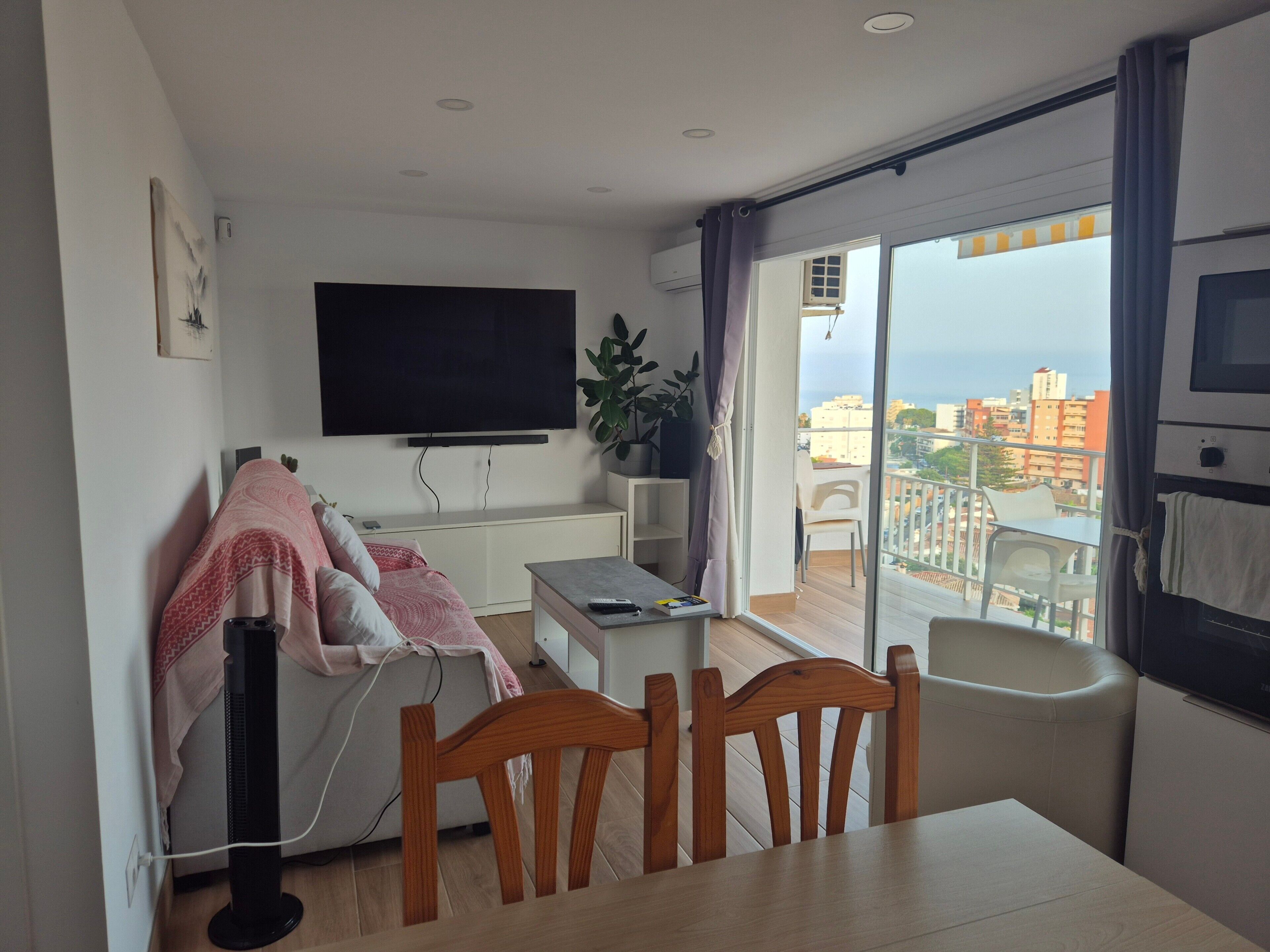 A modern living room with a flat-screen TV, a dining table with chairs, a sofa with a blanket, and a balcony with a view of the city.