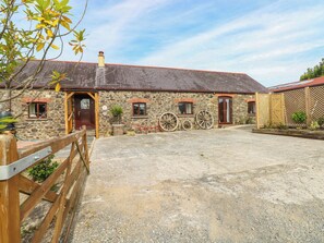 A stone building with a red roof, wooden door, and windows. There are large wheels mounted on the wall and a wooden fence in the foreground. - Coedllys Uchaf Cottage (Carmarthen)