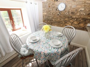 A dining area with a round table set for four, chairs, a clock on the wall, and a window with curtains. - Coedllys Uchaf Cottage (Carmarthen)