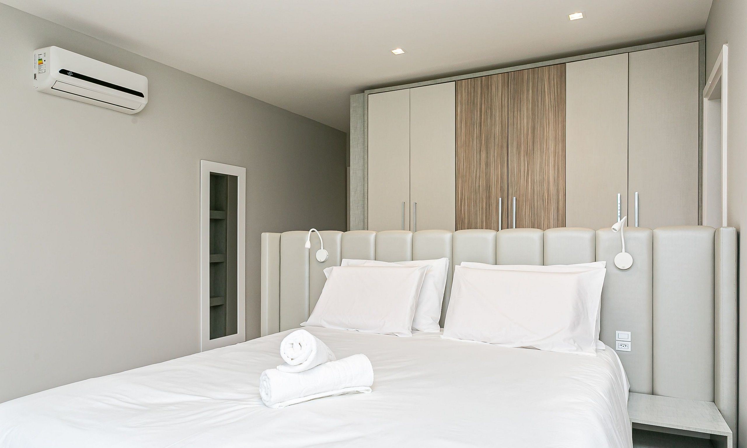 Un dormitorio moderno con cama, ropa de cama blanca, armario empotrado y unidad de aire acondicionado.