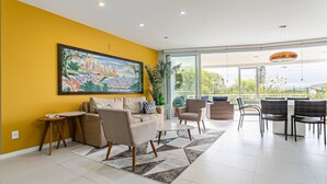 A modern living room with a yellow wall, a large painting, a dining area with chairs and a table, and a view through large windows. - Ap in Resort 200m From the Beach Floripa-sc Mhe003 (Florianópolis)