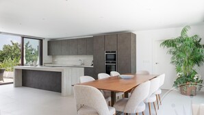A modern kitchen with a central island, a dining area with a wooden table and chairs, and a large potted plant. - La Manga Club Individual Villa 619 (Atamaría)