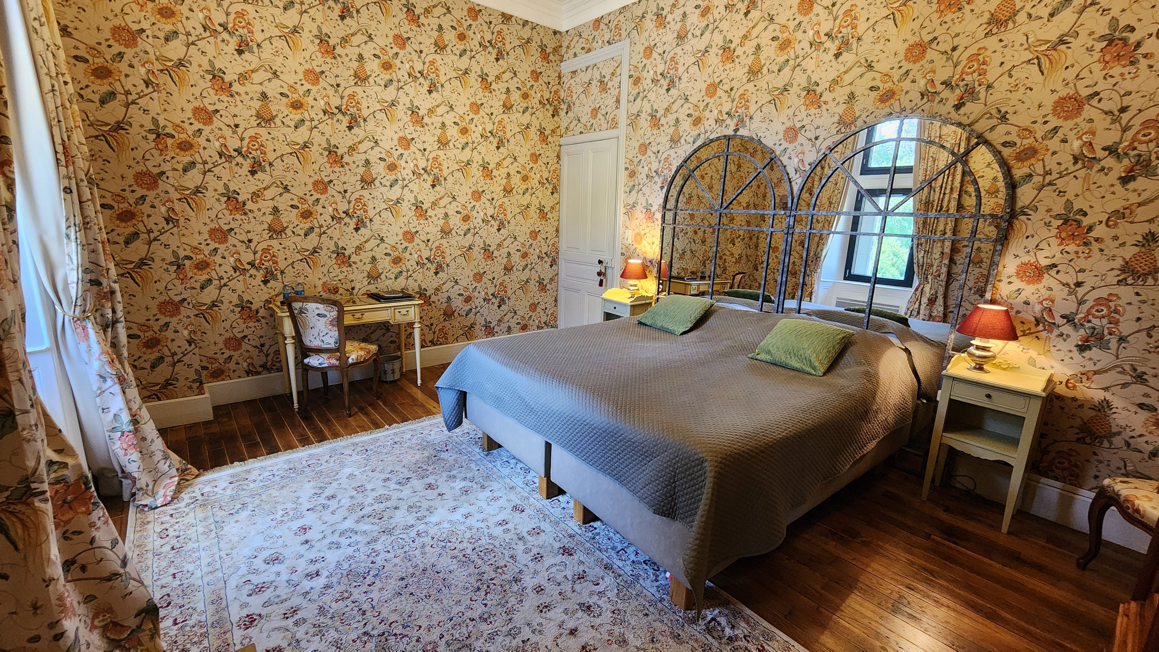 Une chambre avec un papier peint à motifs floraux, un lit avec des coussins verts, une table de chevet avec une lampe et un bureau en bois avec une chaise.