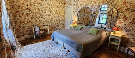A bedroom with floral wallpaper, a bed with green pillows, a nightstand with a lamp, and a wooden desk with a chair.