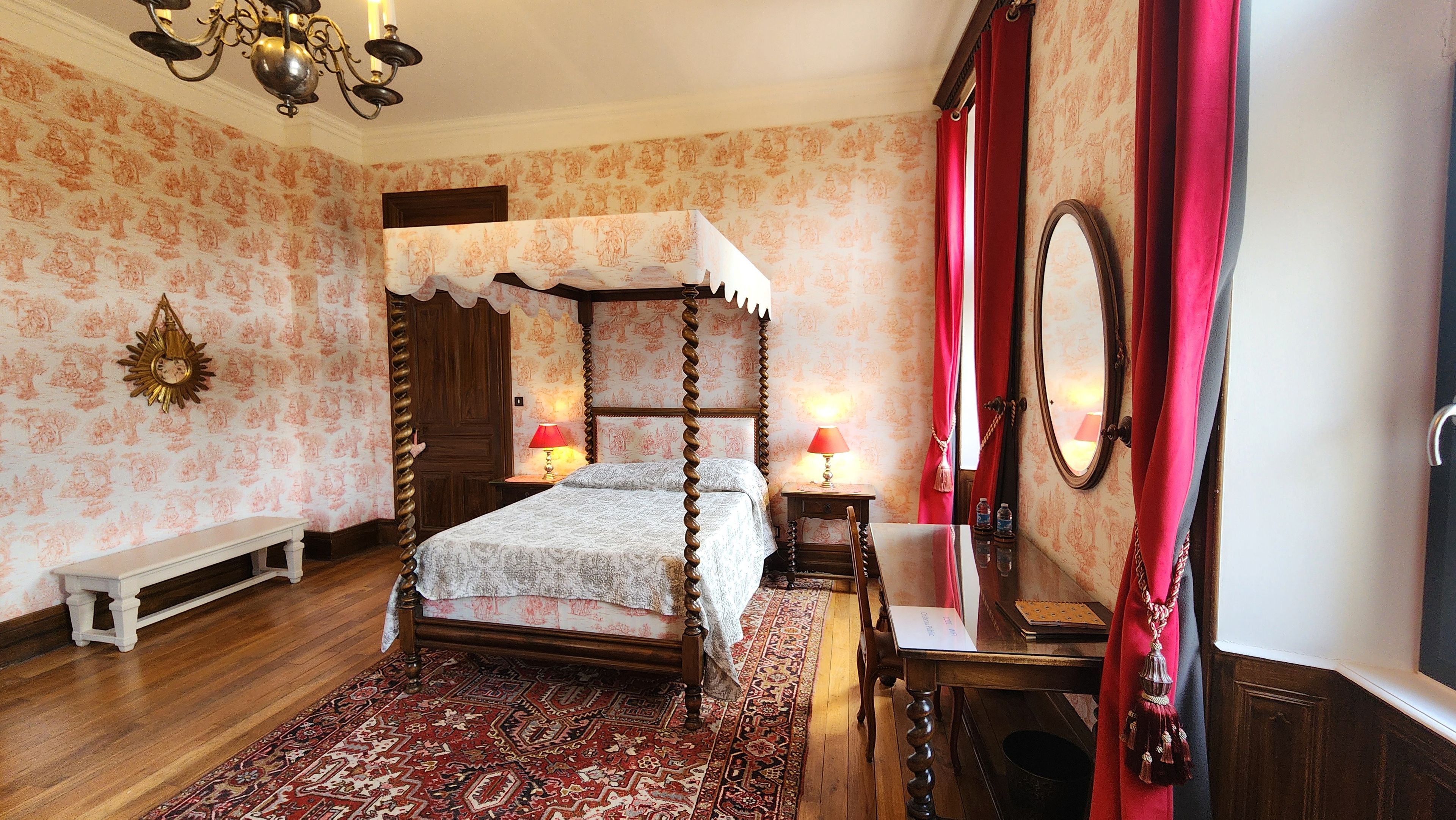 Een traditioneel hemelbed met gordijnen, een houten bankje, een bureau met spiegel, en een kamer met behang met bloemenmotieven en een kamerbreed tapijt.