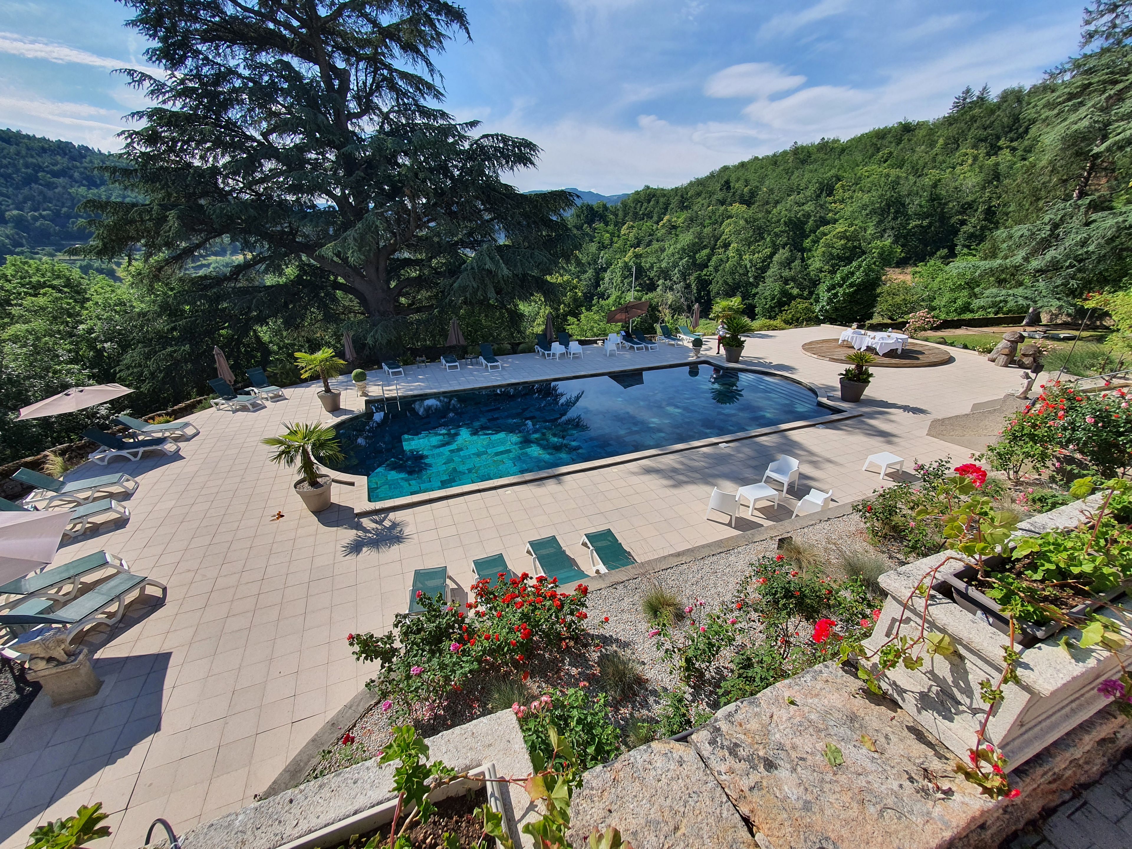 Une piscine entourée de chaises longues et d’une table dressée pour les repas, le tout baigné de verdure et surplombé par un paysage montagneux.