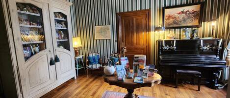 Een kamer met een piano, een houten kast met glazen deuren, een tafel met tijdschriften en gestreept behang.