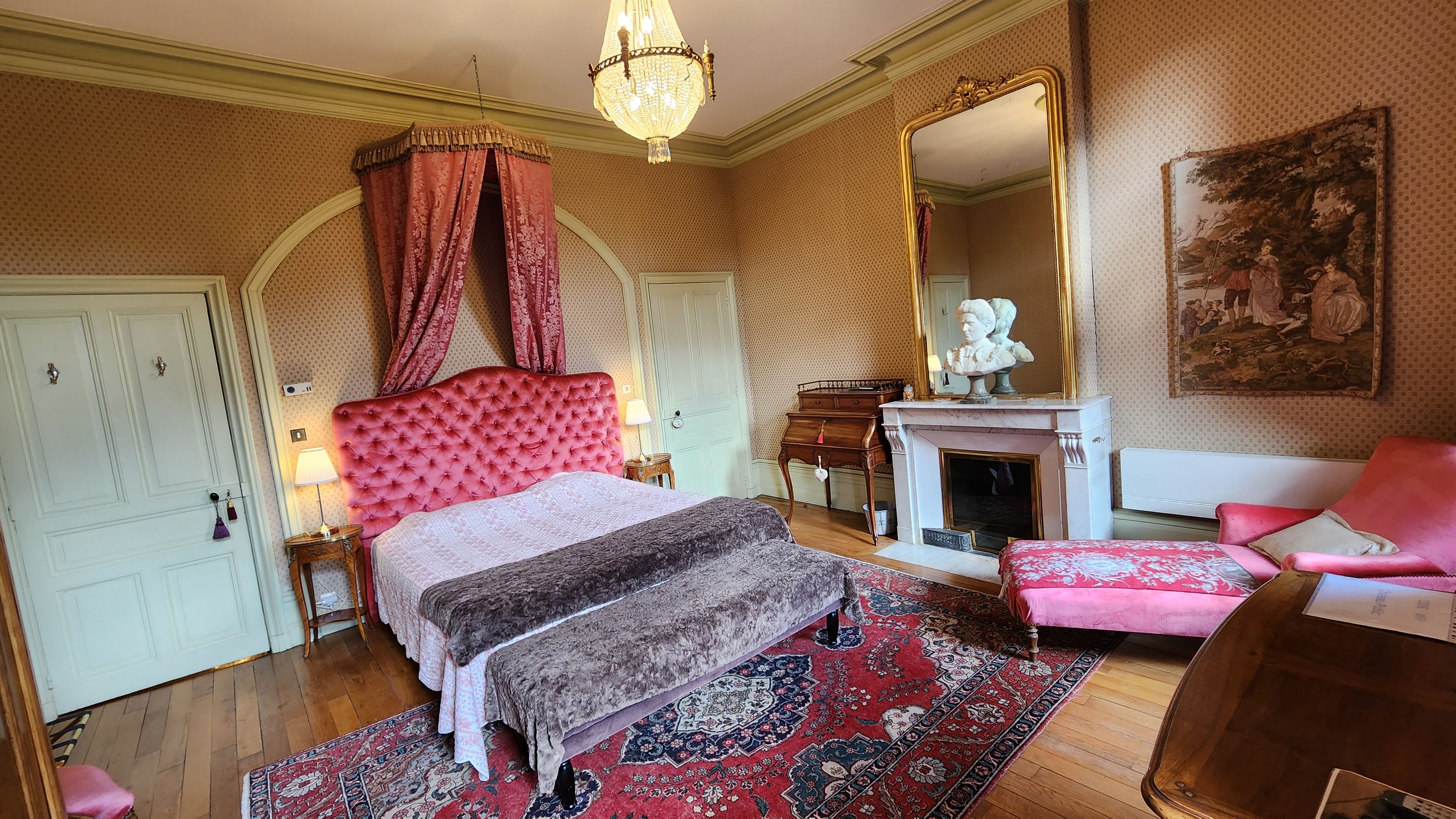 Een slaapkamer met een groot bed, een open haard, een spiegel en een rode stoel.