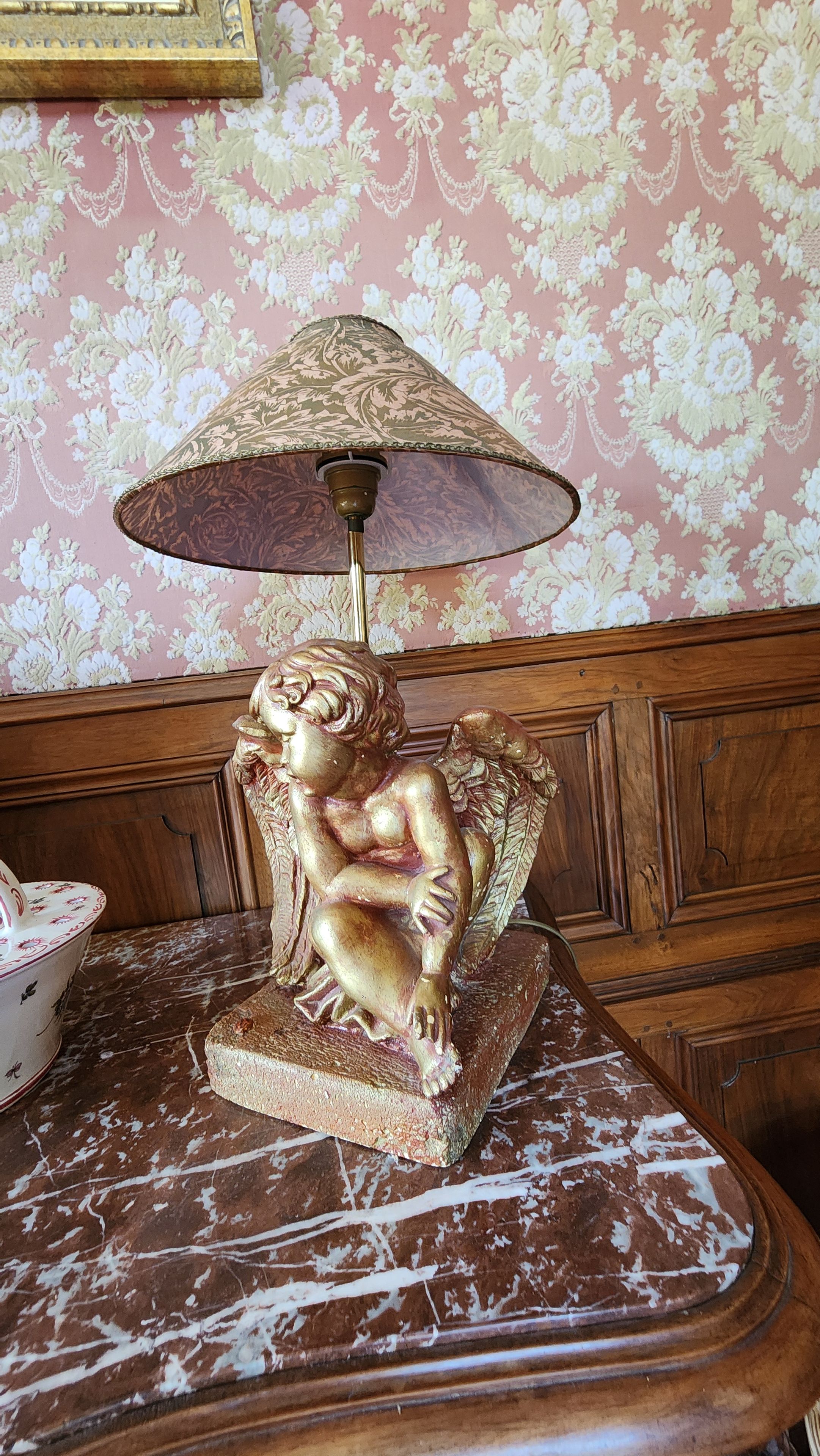 A table with a decorative lamp featuring a golden statue of a seated figure and a patterned lampshade.