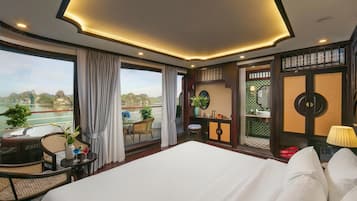 A hotel room with a large bed, a balcony with a view of the water, a small table with a plant, and a bathroom with a mirror and sink.