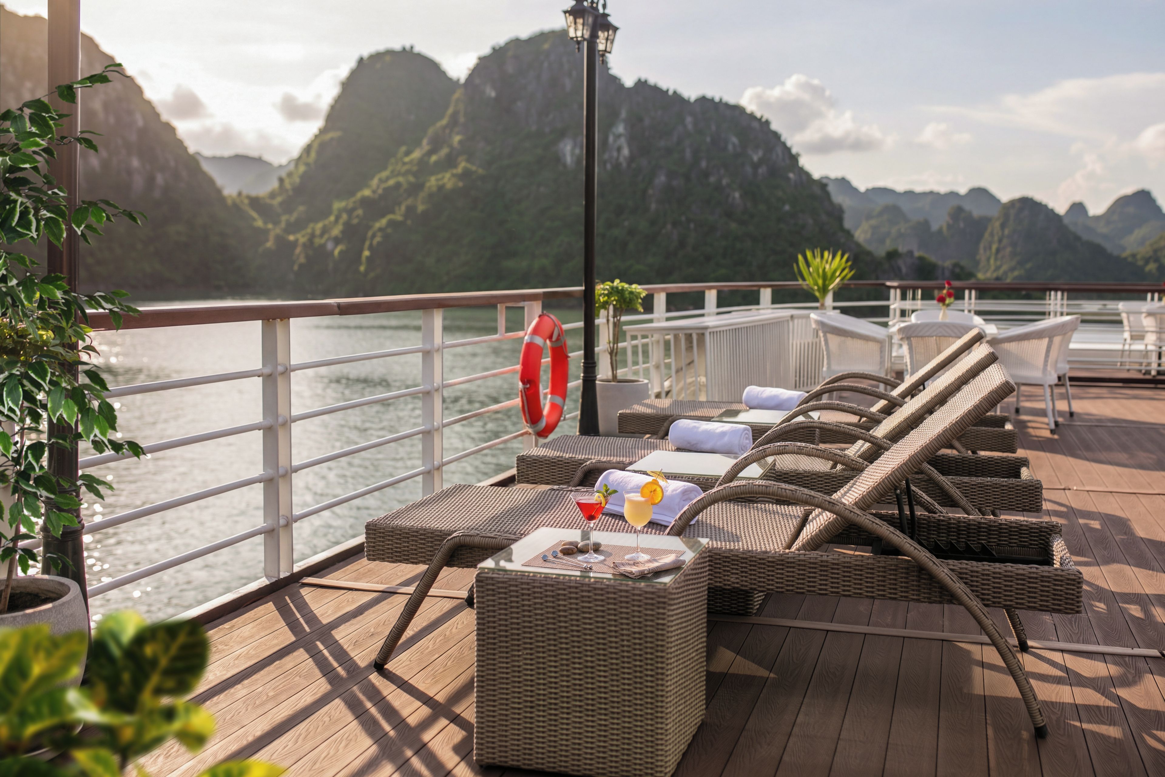 A deck with wicker furniture, a lifebuoy, and a drink on a table with a view of mountains.