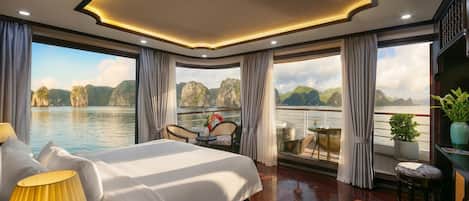 A spacious bedroom with a large bed, a seating area with a table, and a view of a body of water with mountains.