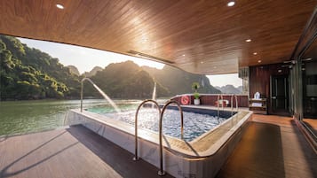 A boat's interior with a hot tub, wooden deck, and a view of a forested coastline.