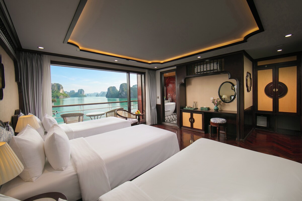 A hotel room with two beds, a balcony with a view of the sea, a bathroom, and a small sitting area.