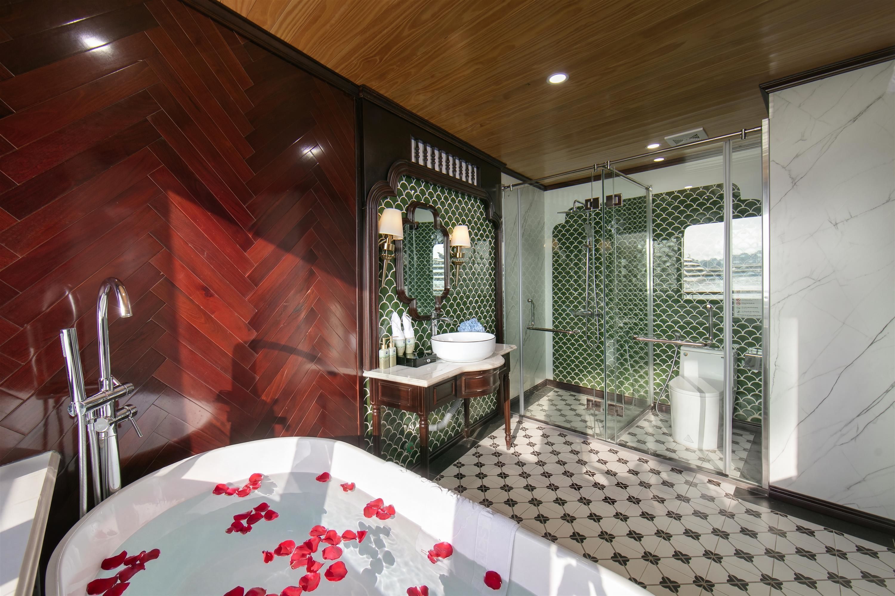 A modern bathroom with a herringbone wood floor, a freestanding bathtub with rose petals, a glass-enclosed shower, and a vanity with a green patterned backsplash.