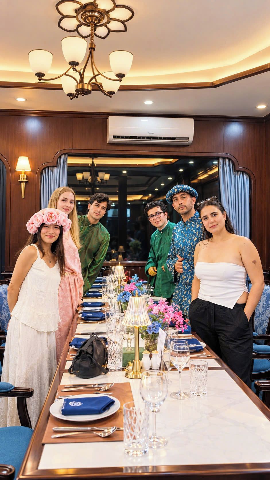 A group of people seated at a long dining table with a chandelier above, in a room with wooden paneling and an air conditioning unit on the wall.