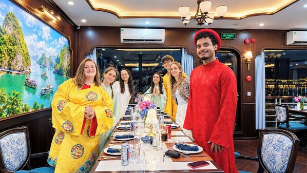 A group of people seated at a table in a room with a large scenic painting on the wall. - Hermes Premium Cruise (Hong Gai)