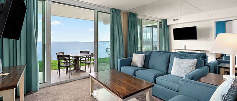 A hotel room with a sofa, a coffee table, a TV, and a view of the sea.