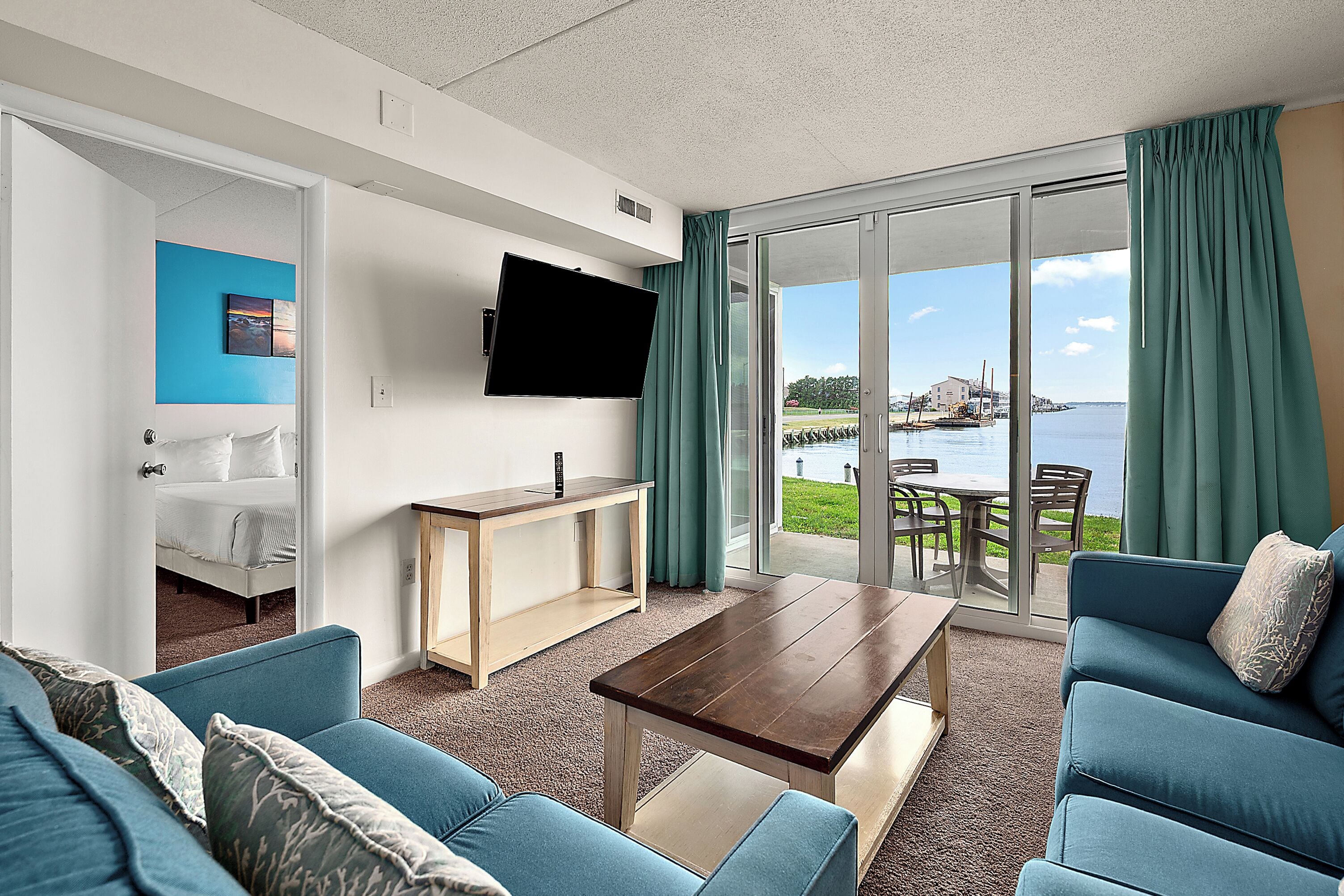 A modern living room with a sofa, a coffee table, a TV mounted on the wall, and a view of the sea through large glass doors.